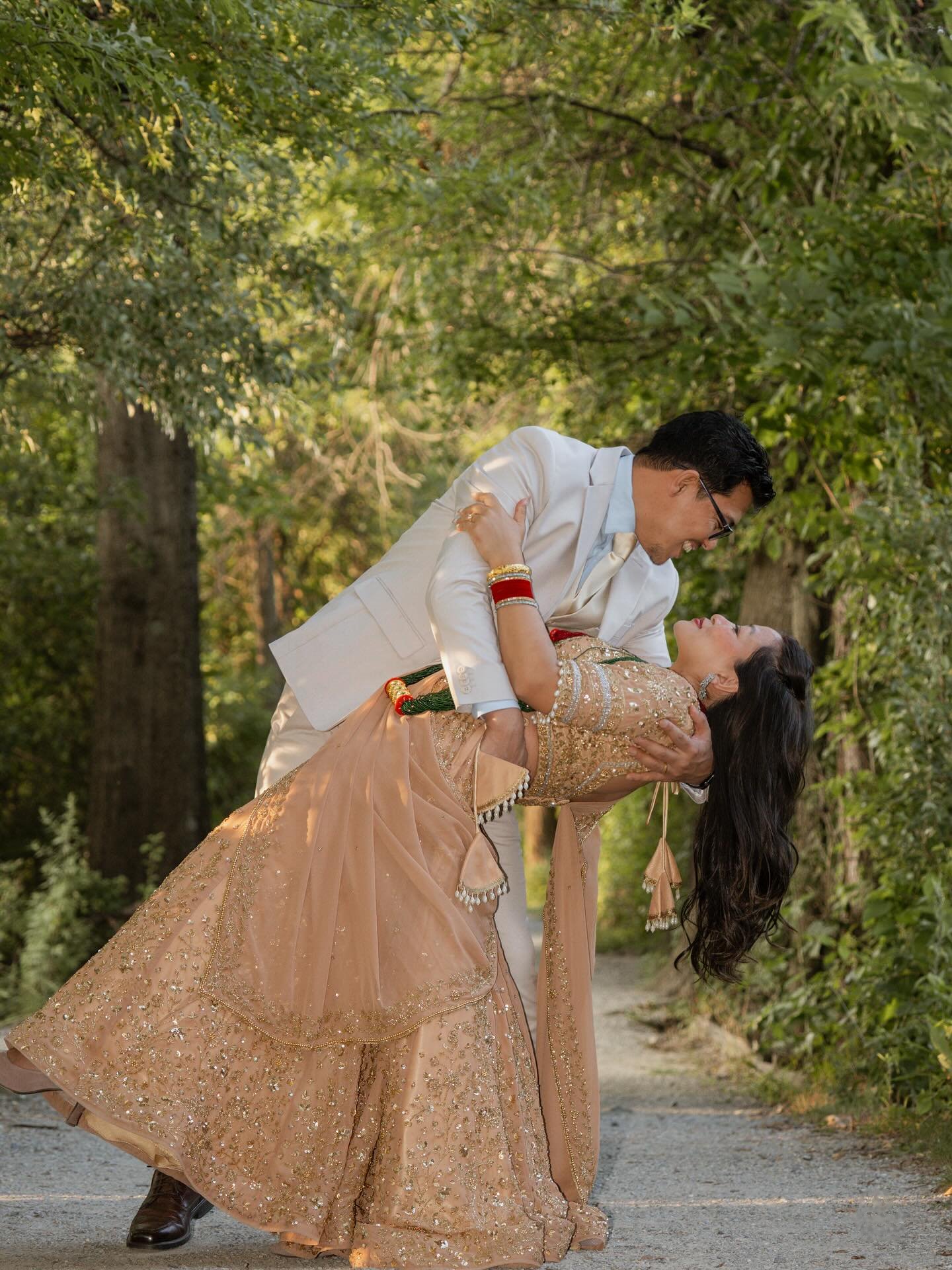 A man in a cream suit dipping a woman in a gold lehenga on a tree-lined path