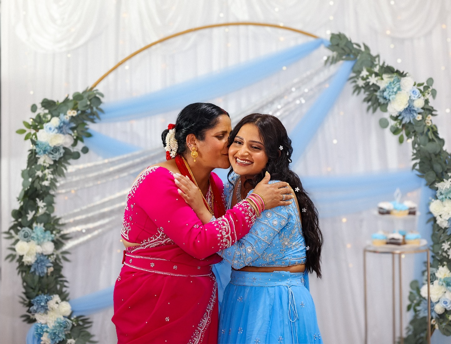 A mother kissing her daughter's cheek in front of a blue and white floral arch