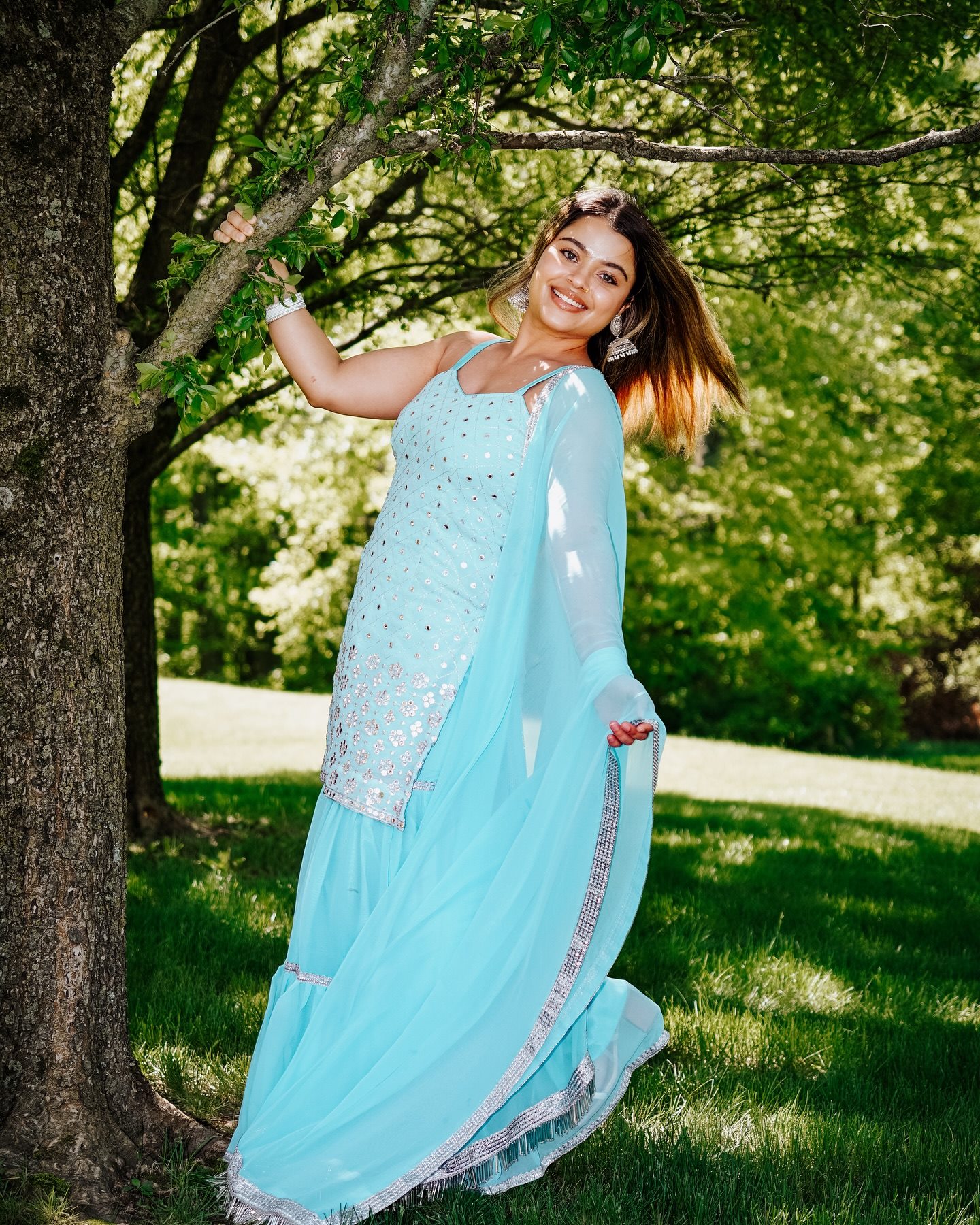 A woman in a light blue outfit swinging around a tree, laughing