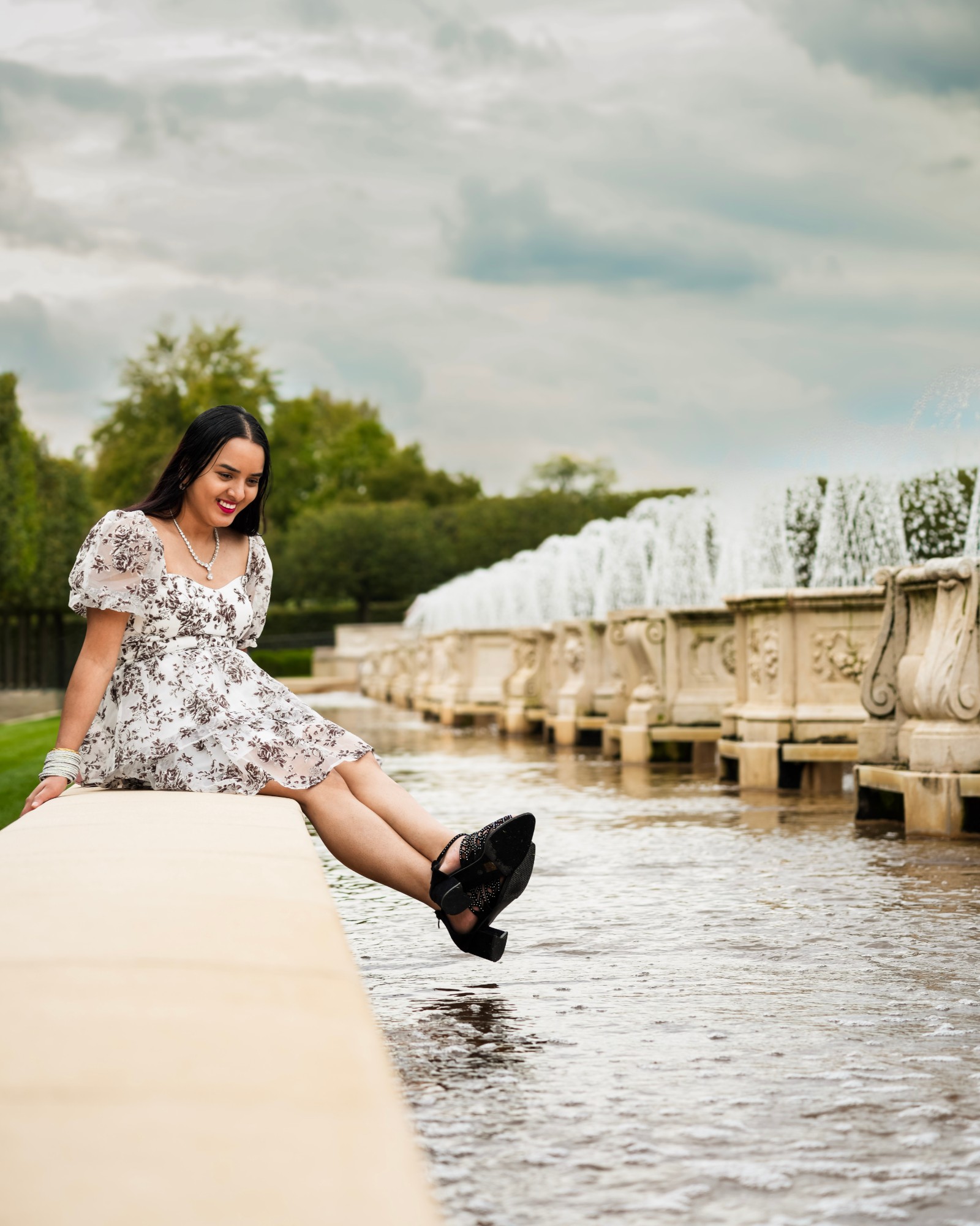 A woman in a floral dress sitting on the edge of a fountain pool, laughing down at the water