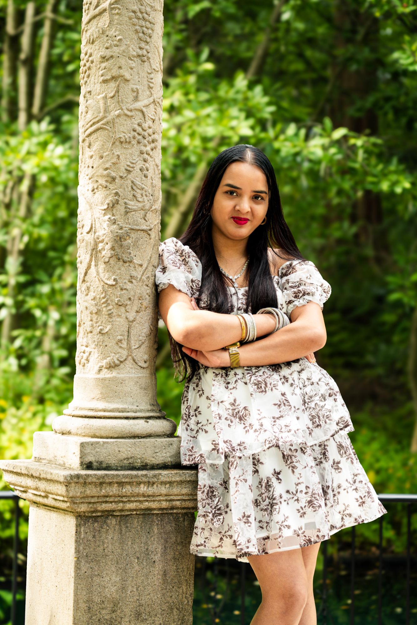 A woman in a floral dress leaning against a carved stone column with her arms crossed