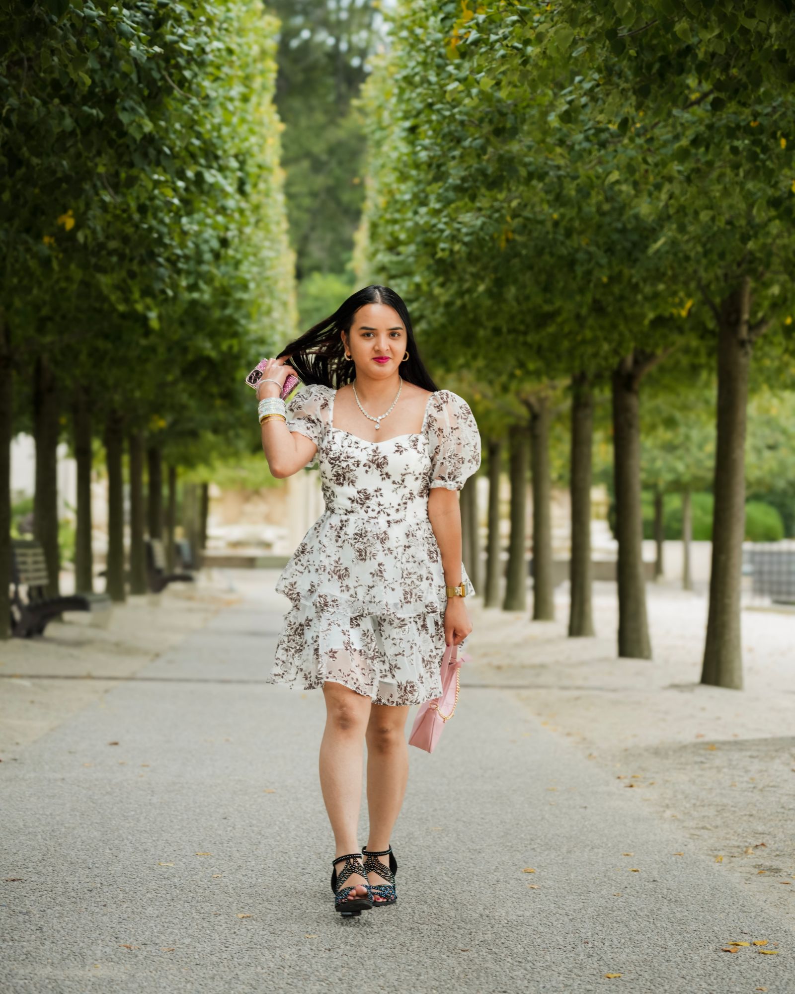 A woman in a floral dress walking down a tree-lined path, one hand in her hair