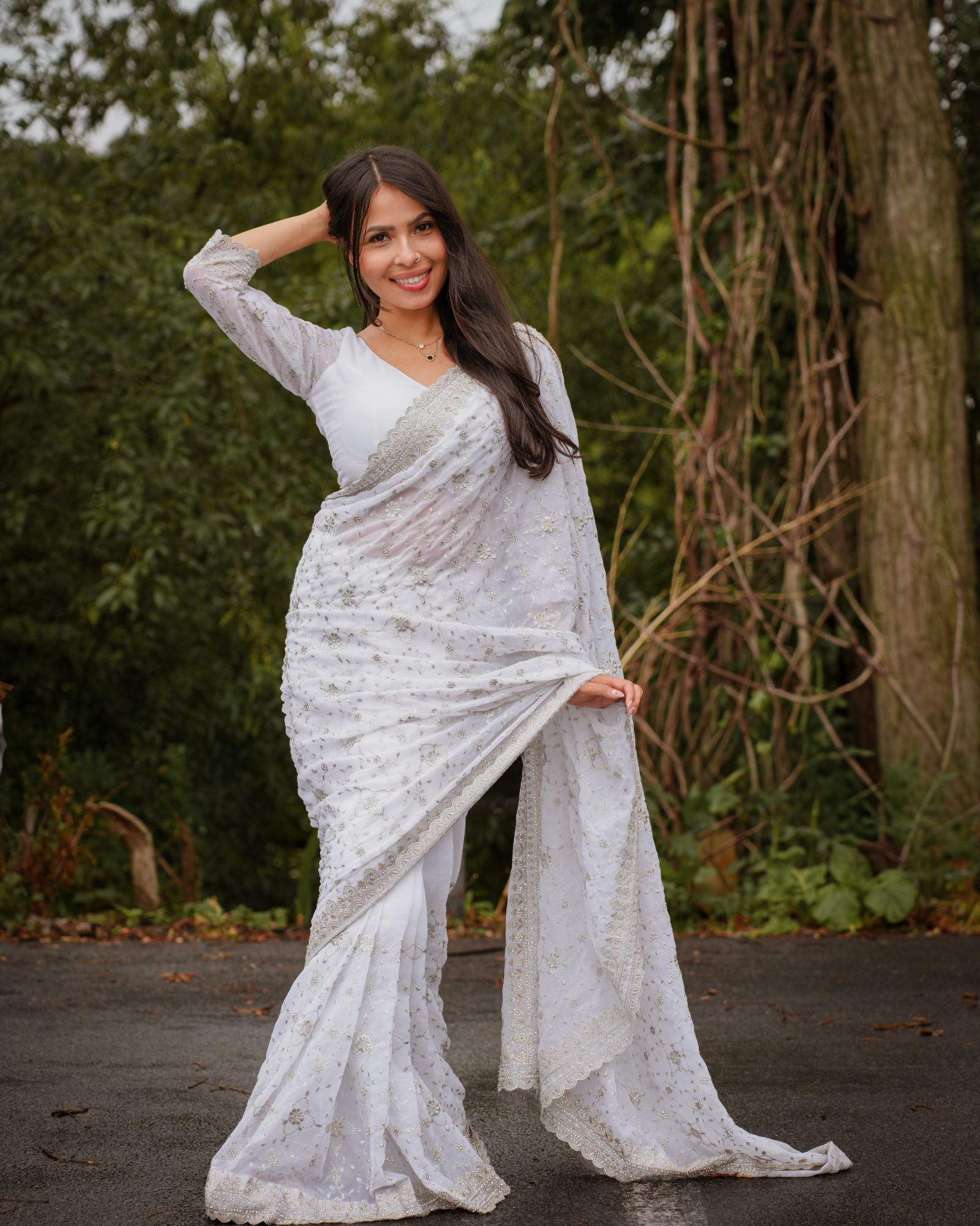 A woman in an embroidered white saree smiling with one hand in her hair