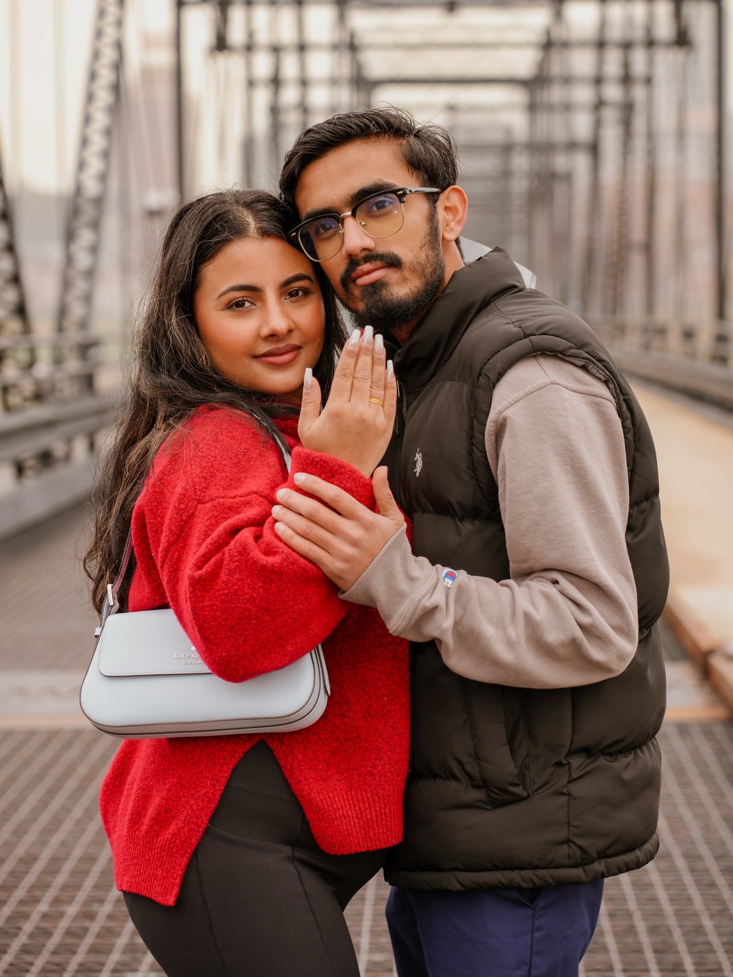 A newly engaged couple on an iron bridge, she holds up her hand to show the ring