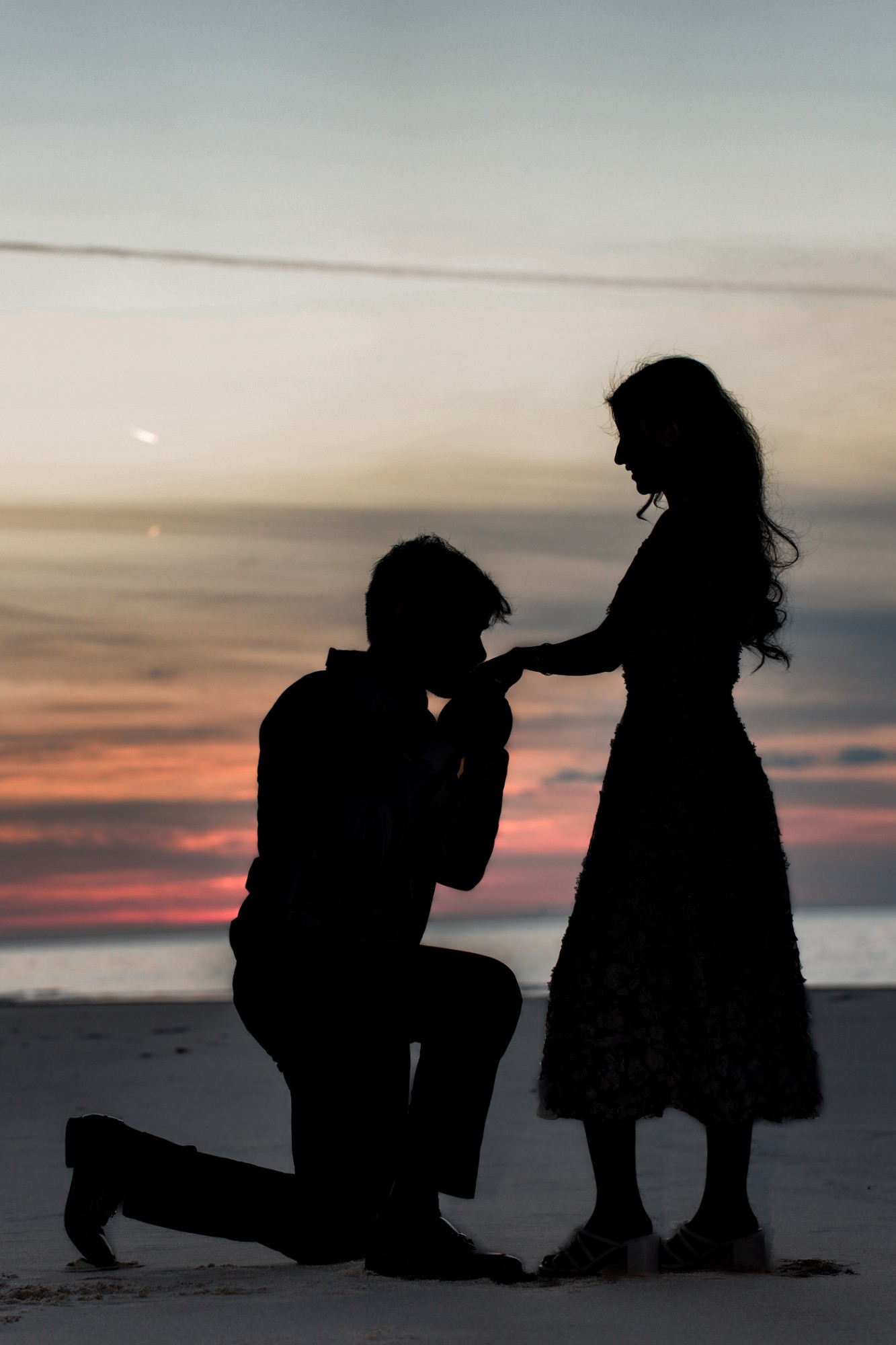 A silhouette of a man on one knee kissing a woman's hand on the beach at sunset