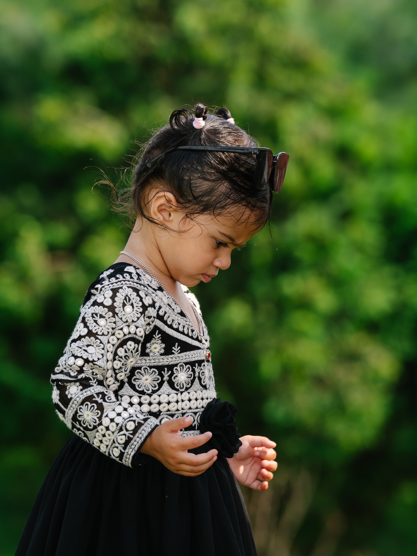 A toddler in an embroidered black dress looking down, sunglasses pushed up on her head
