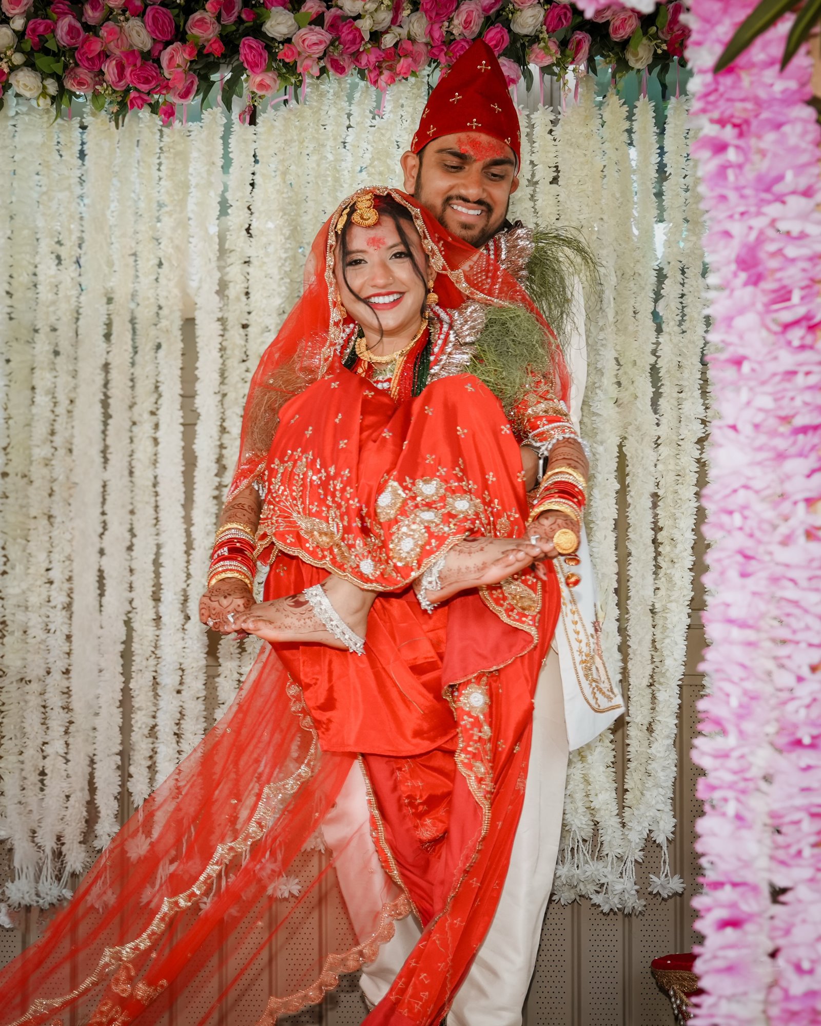 A groom in a red topi holding his bride from behind, both smiling, her red veil draped around them