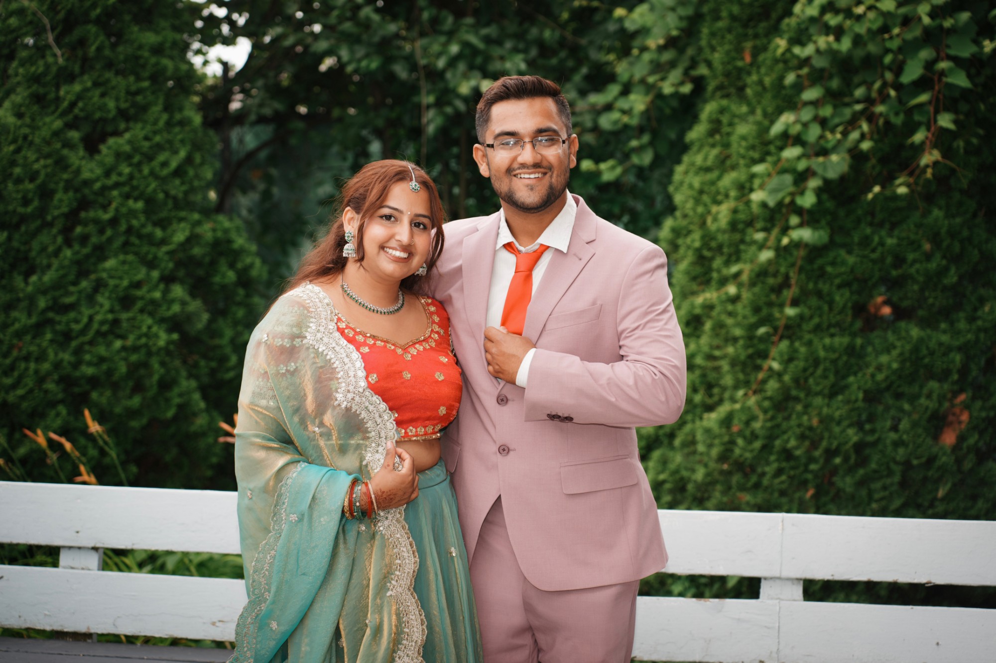 A couple in a lehenga and a blush-pink suit smiling in front of garden hedges