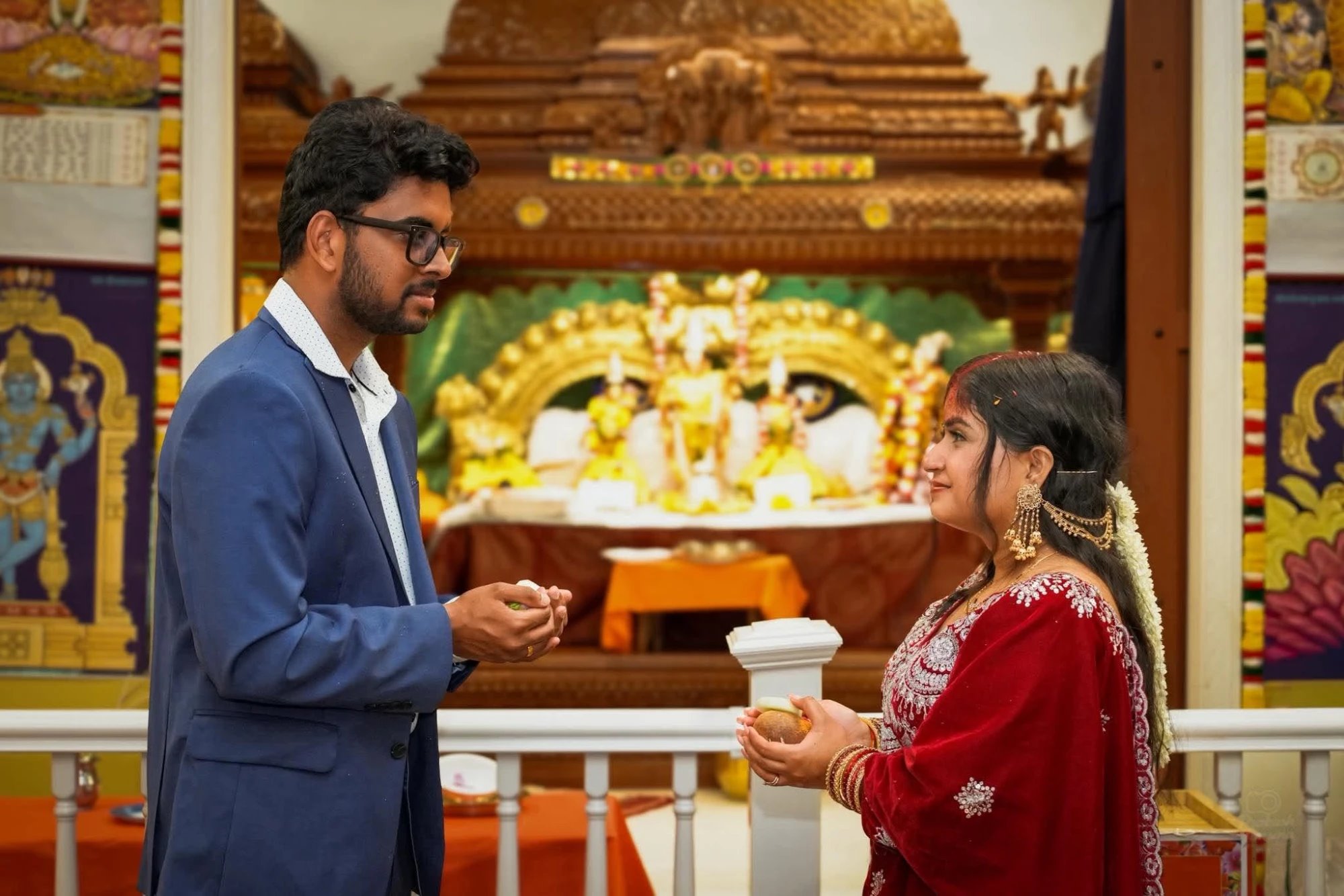 A bride and groom facing each other inside a temple, each holding offerings