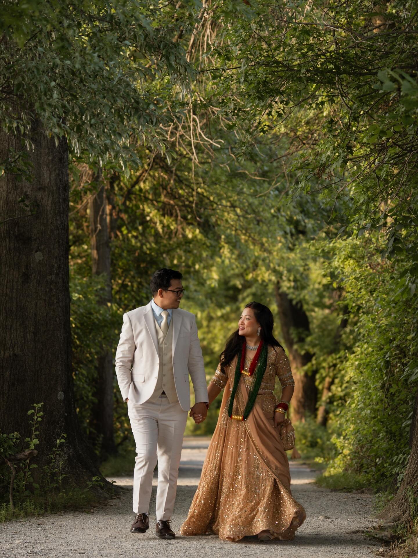 A couple in wedding clothes walking hand in hand down a tree-lined path, smiling at each other